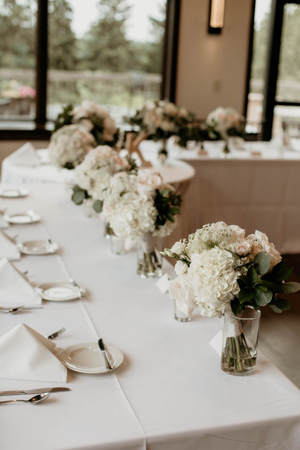 Long white head table decorated with white hydrangea and blush rose floral arrangements at wedding.