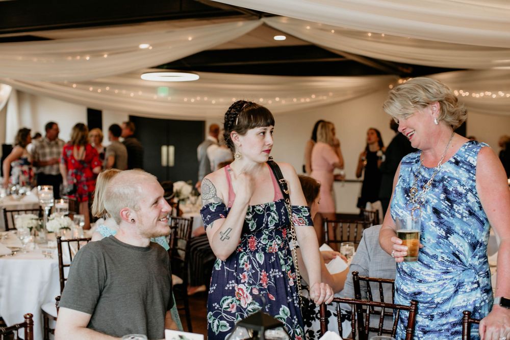 Wedding guests mingle and converse in decorated reception hall with draped ceiling and string lights.