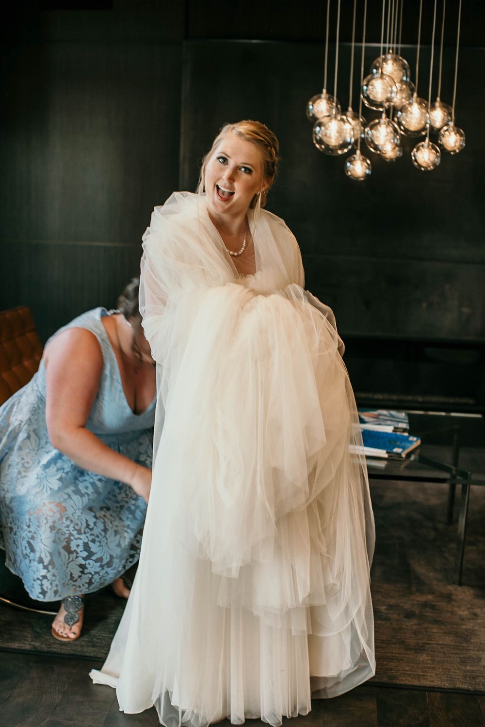 Excited bride in a flowing white tulle gown being helped by a bridesmaid before the wedding.
