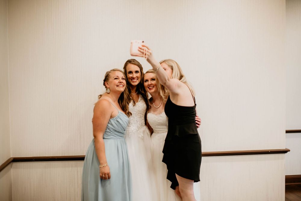 Bride in white gown takes selfie with two guests in light blue and black dresses at wedding reception.