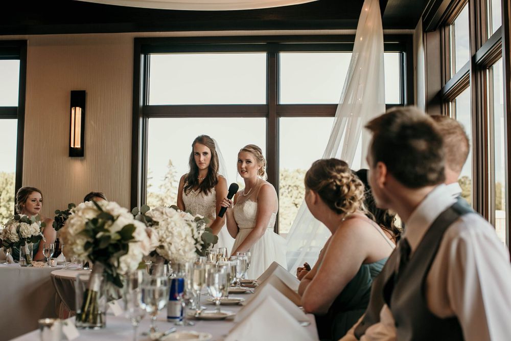Two brides stand at wedding reception head table giving a speech with microphone amid white floral centerpieces.