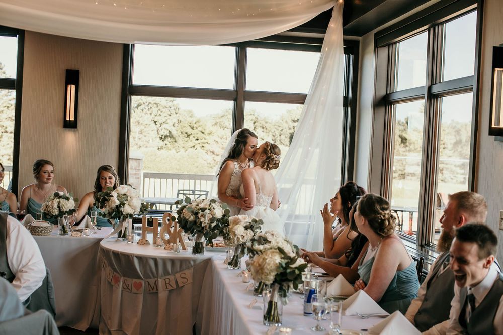 Two brides share a kiss at wedding reception head table with guests seated around elegant floral arrangements.