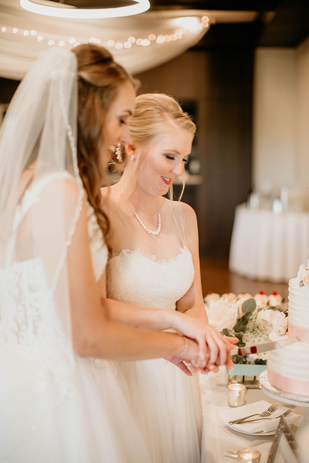 Two brides in white gowns smile while cutting their two-tier wedding cake together at reception.