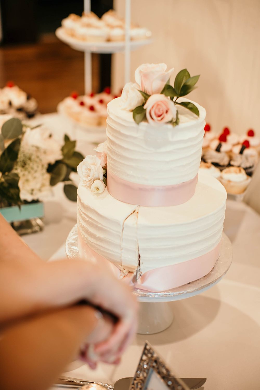 Elegant two-tier white wedding cake with pink ribbon and rose topper being cut at reception dessert table.