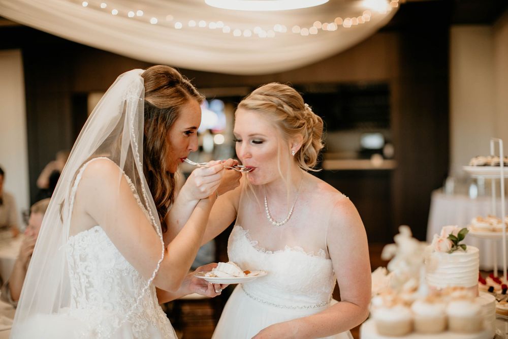 Bride in veil feeds wedding cake to her bride in pearl necklace at elegant reception dessert table.