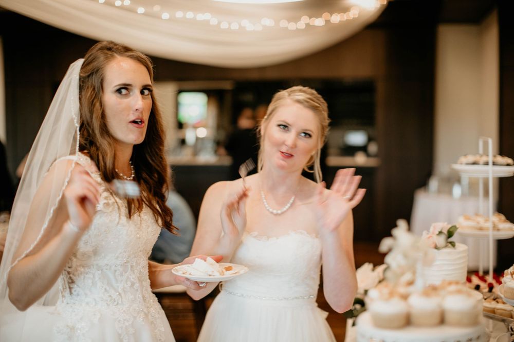 Two brides make funny faces while eating wedding cake near dessert table with cupcake tower at reception.