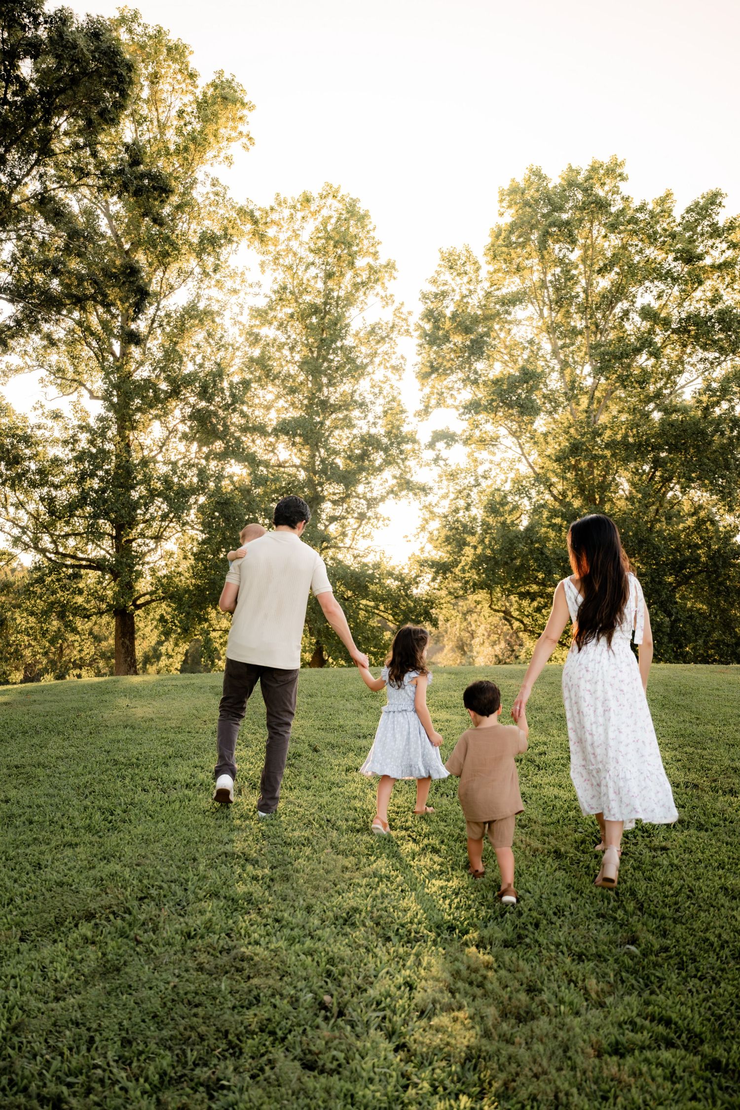 Family of four holding hands walking through a lush green park at golden hour sunset.