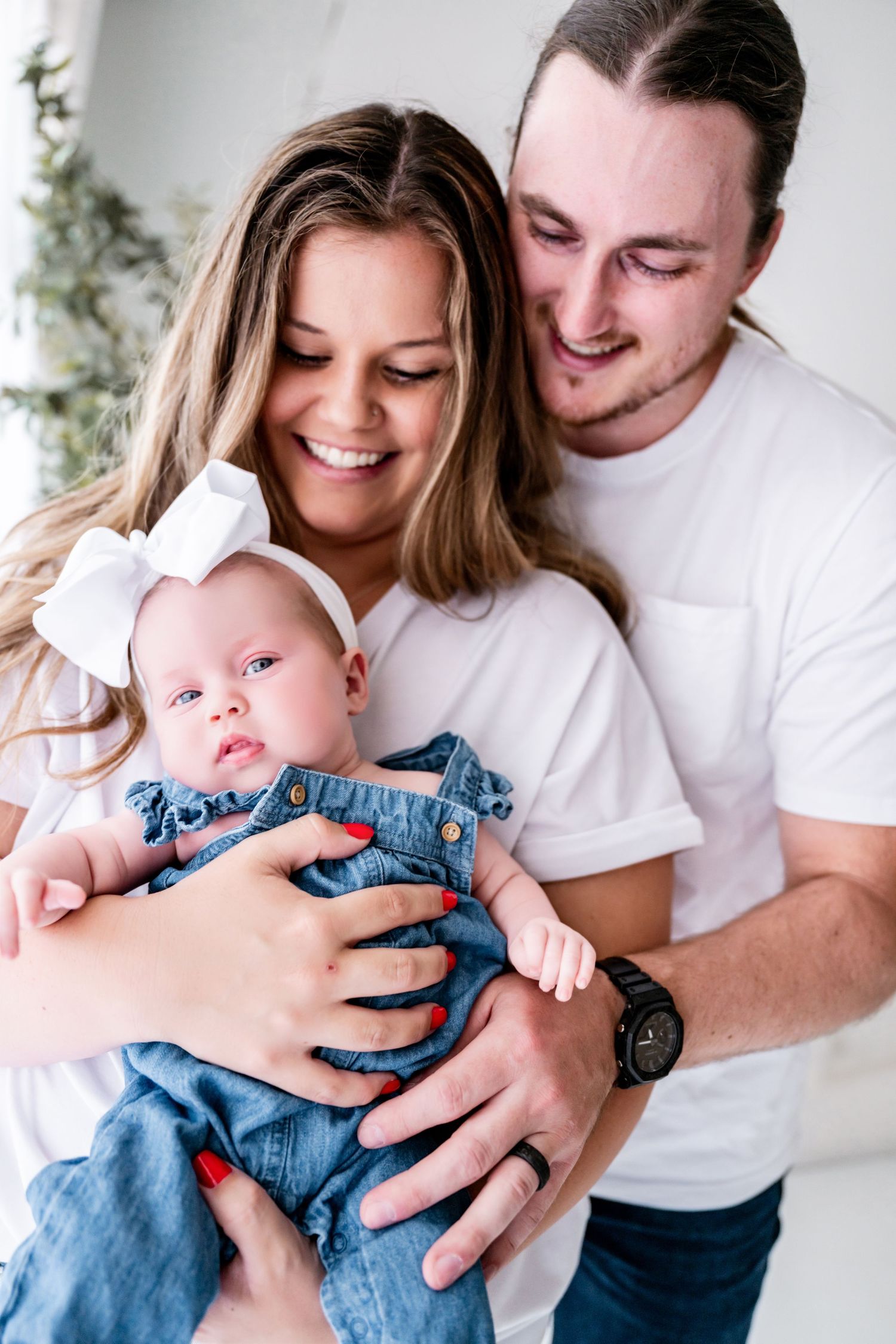 Happy young couple in white t-shirts smiling at their baby girl in denim overalls with a white bow headband.
