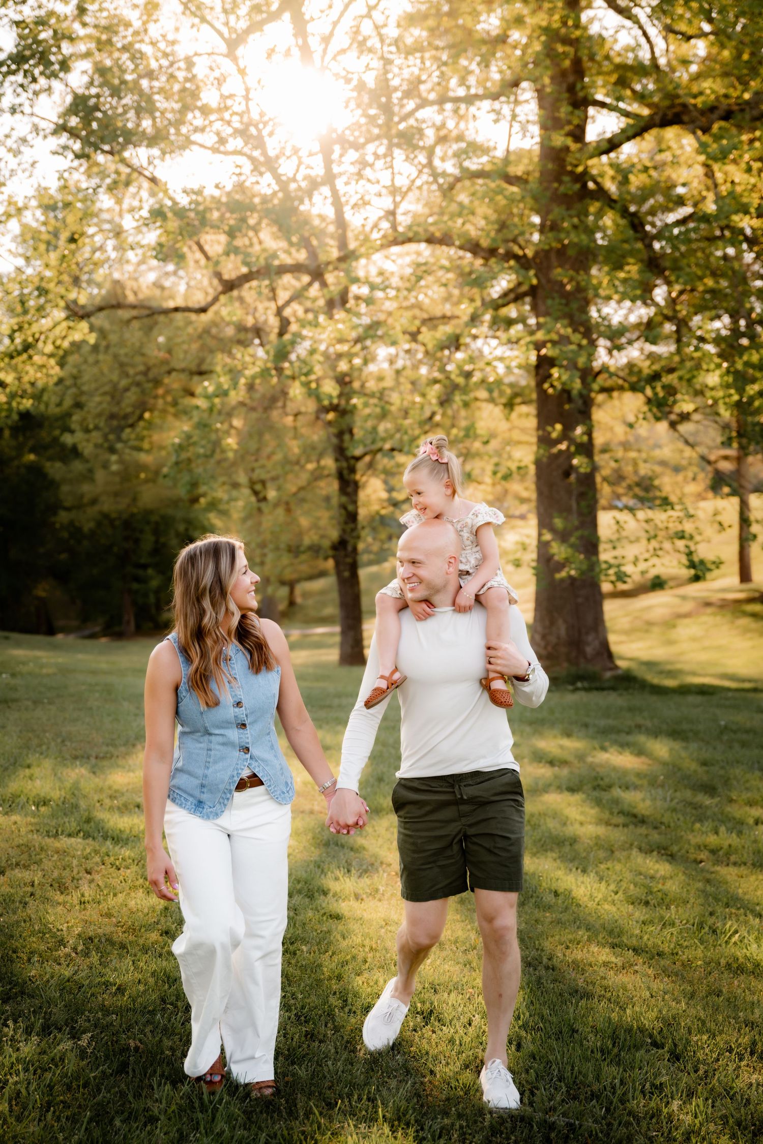 Family walking in sunlit park, man carrying toddler on shoulders, woman in white pants holding hands.
