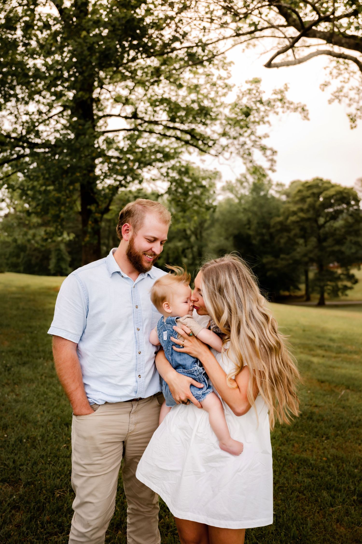 A young family poses outdoors, parents smiling at their baby in a lush green park during golden hour.