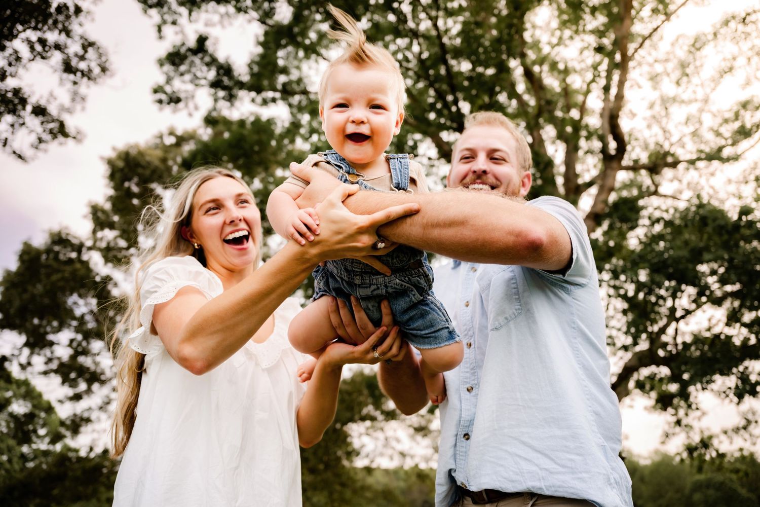 Happy family with laughing parents lifting a joyful baby outdoors among green trees in warm golden light.