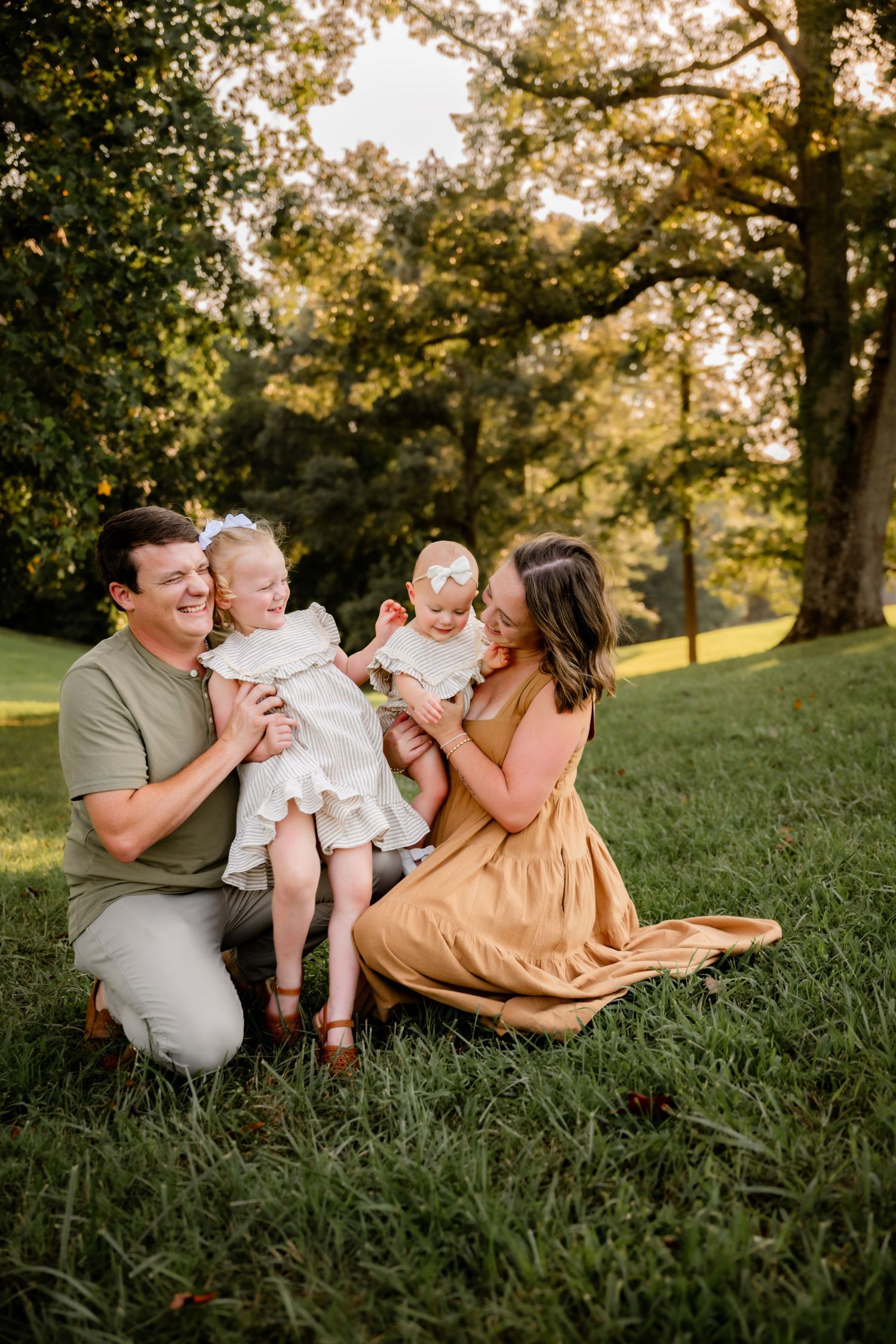 A happy family of four sits on lush green grass, parents smiling at two young daughters in white dresses outdoors.