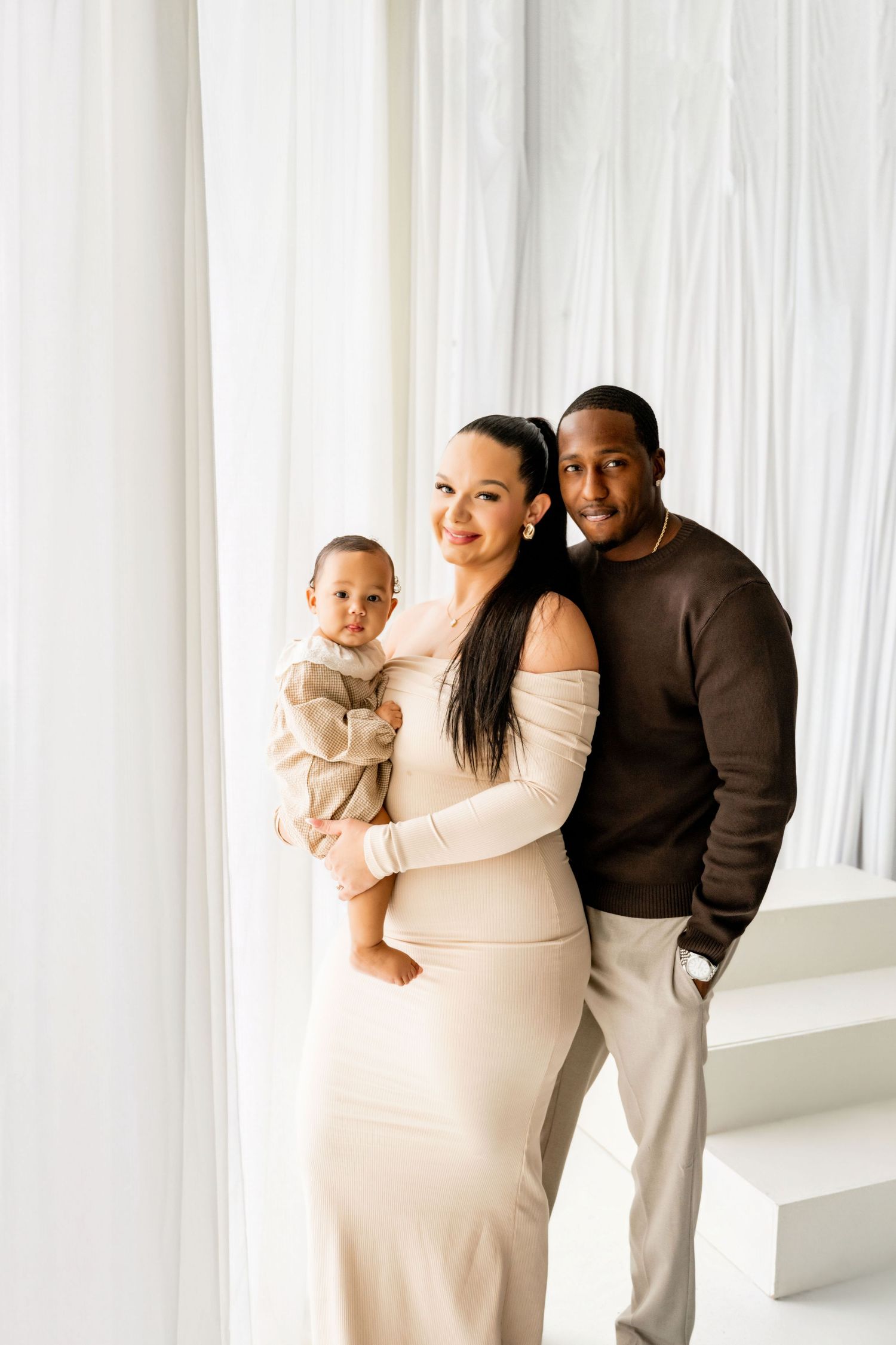 A family of three poses elegantly, mother in cream dress holding toddler, father in brown sweater, against white drape backdrop.
