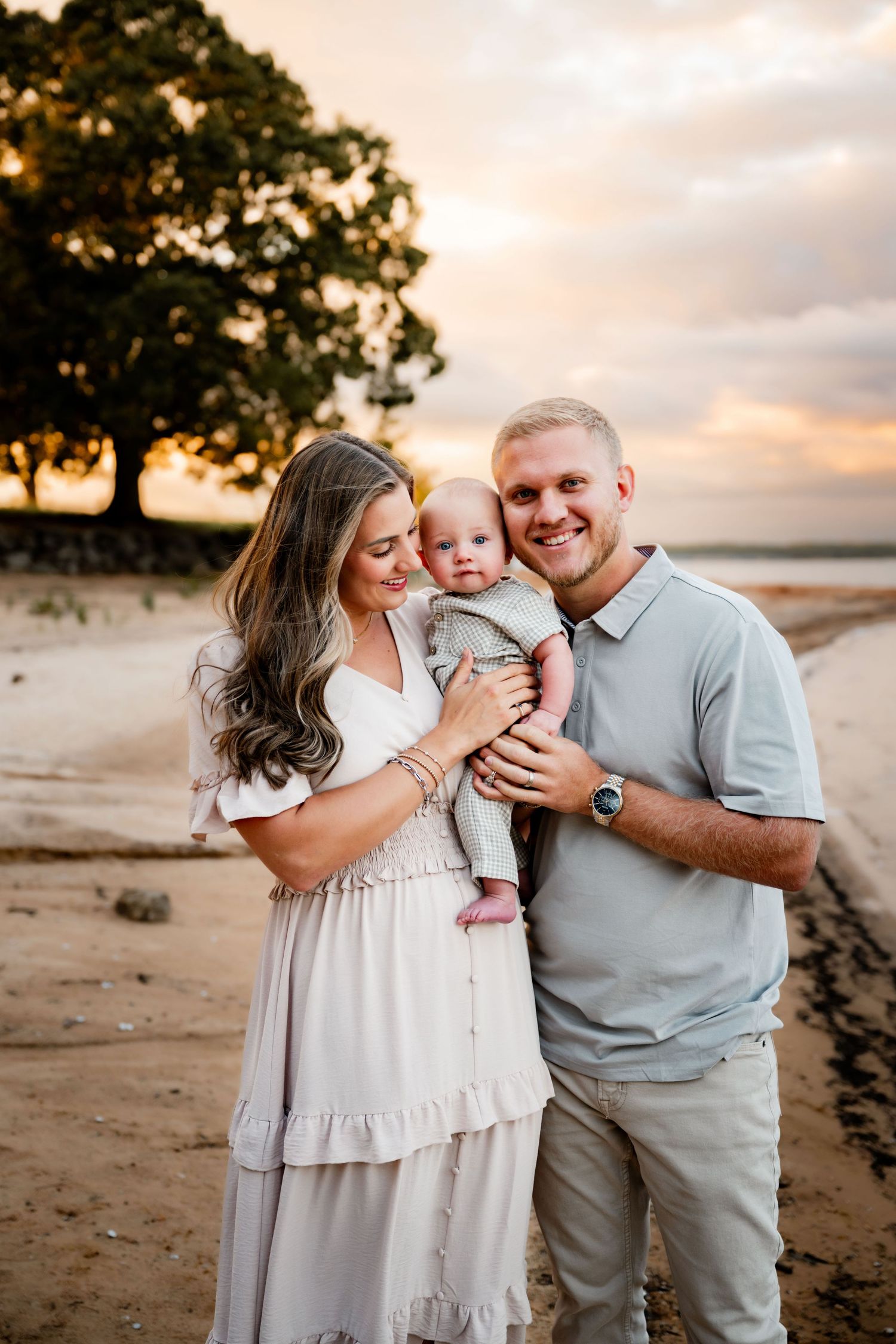 A smiling couple holds their baby on a sandy beach at golden hour with a large tree and colorful sky behind them.