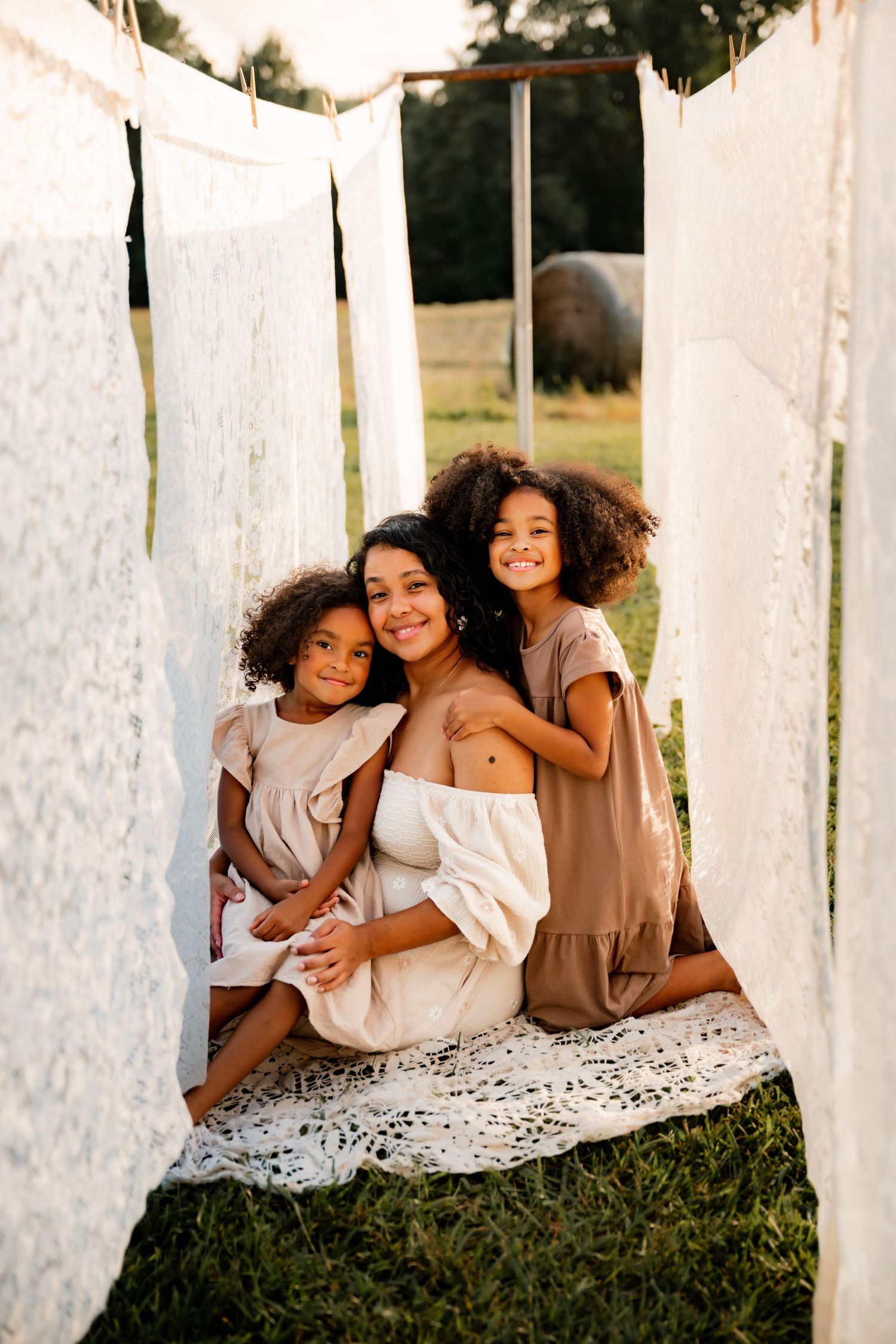 A mother and her two daughters sit together outdoors among hanging white lace fabrics on a green lawn.