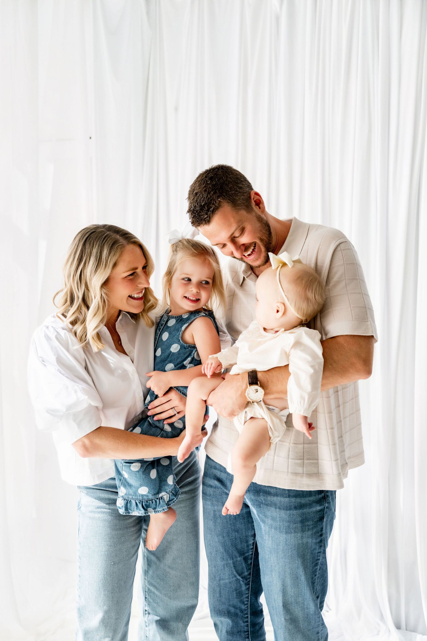 Happy family of four with parents holding toddler girl and baby in white studio setting.