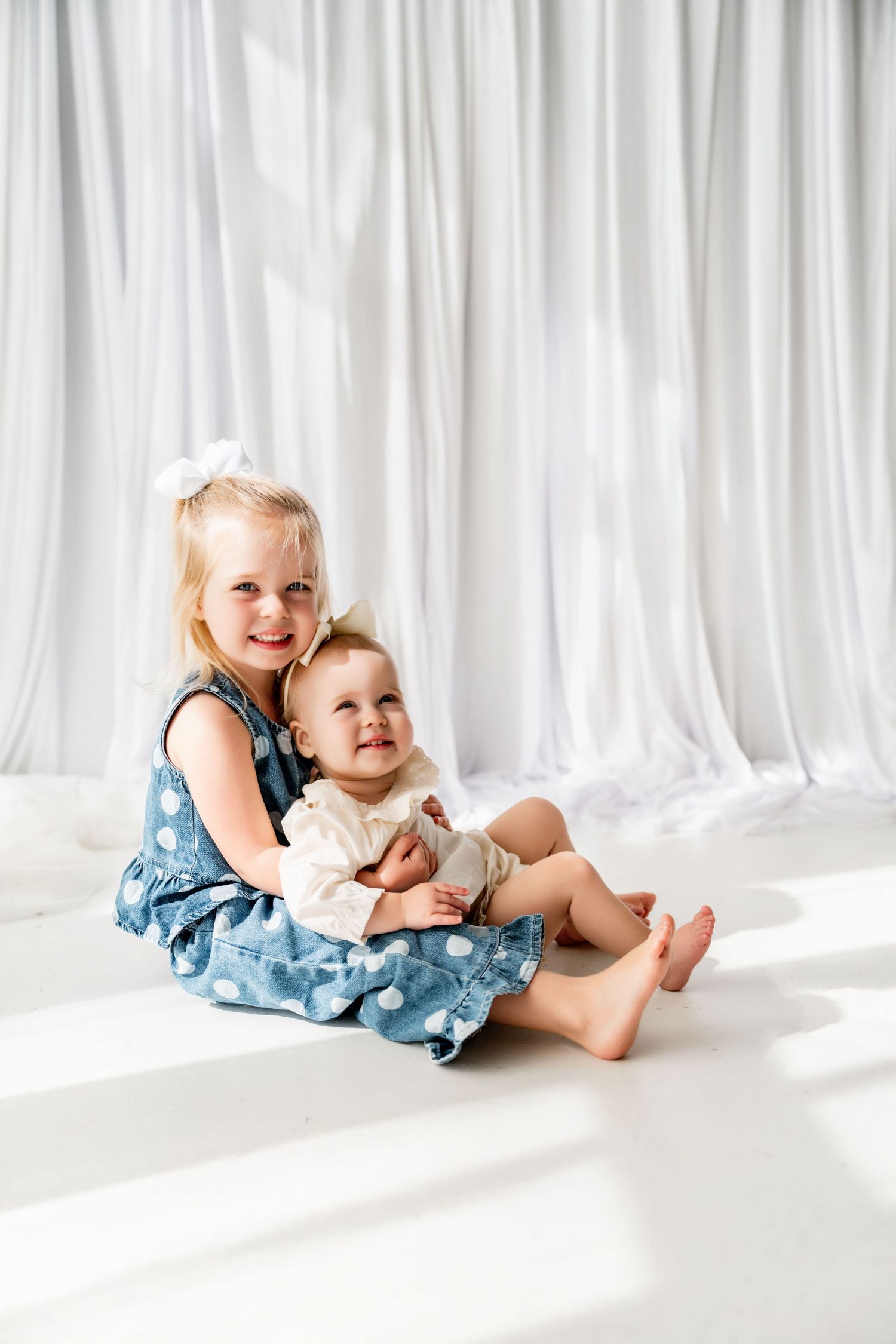 Two young sisters sitting together, older girl in blue polka dot dress hugging smiling baby, white curtain backdrop.