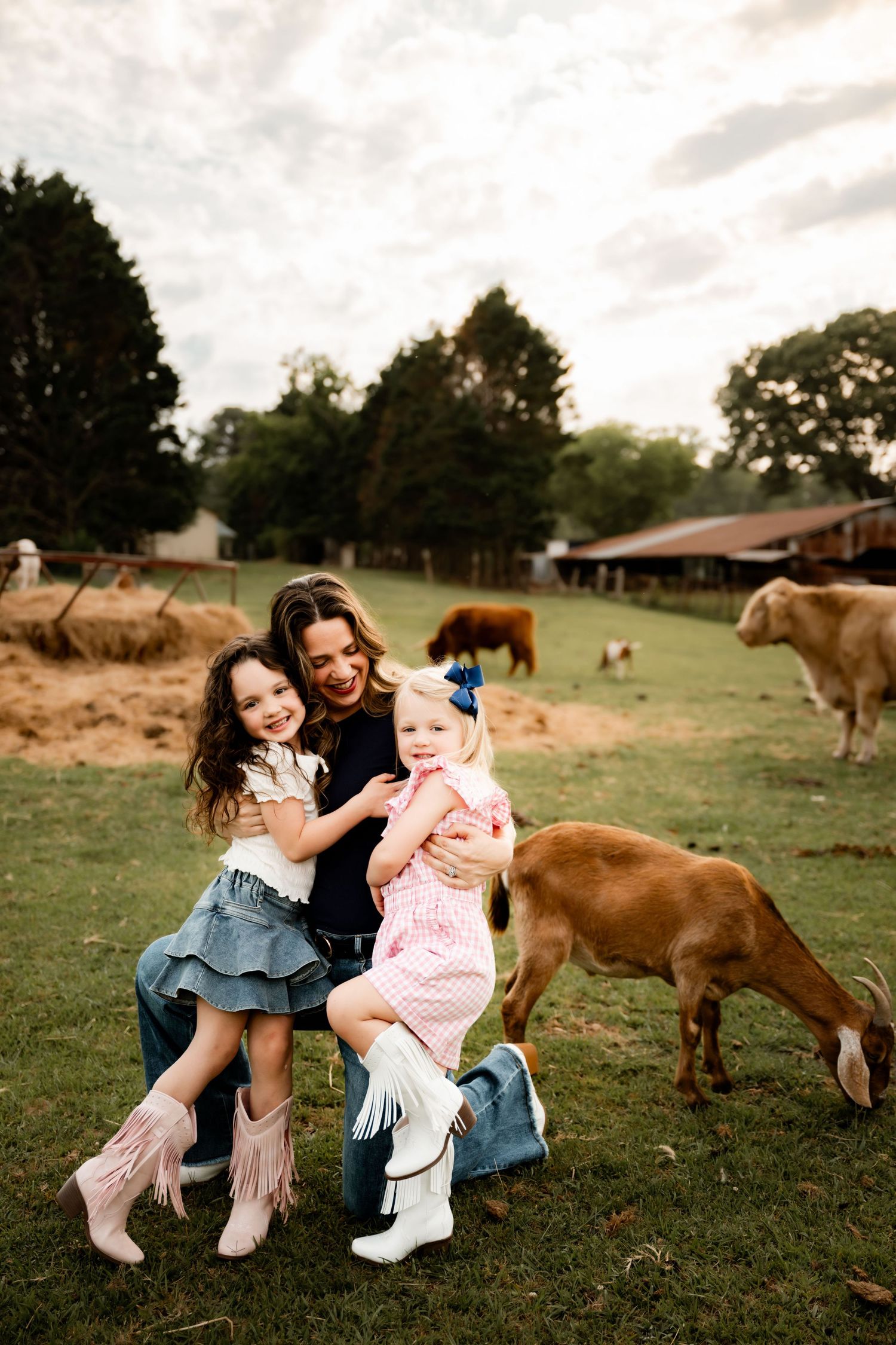 A mother and two young daughters in cowboy boots smile together on a farm with goats and cattle grazing nearby.