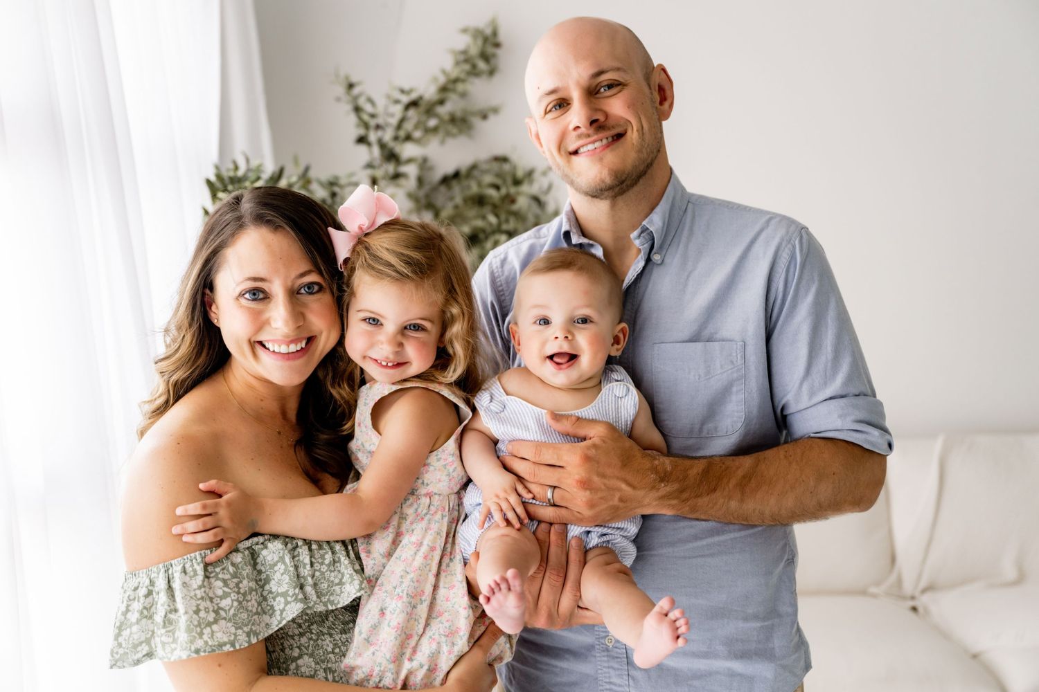 Happy family of four posing together, parents holding two young daughters, smiling in a bright white studio setting.