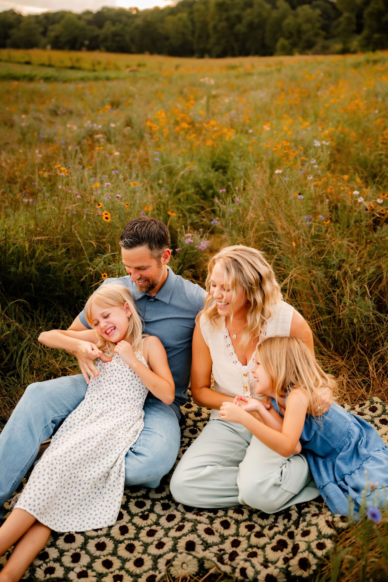 A joyful family of four laughs together on a blanket in a golden wildflower field during sunset.