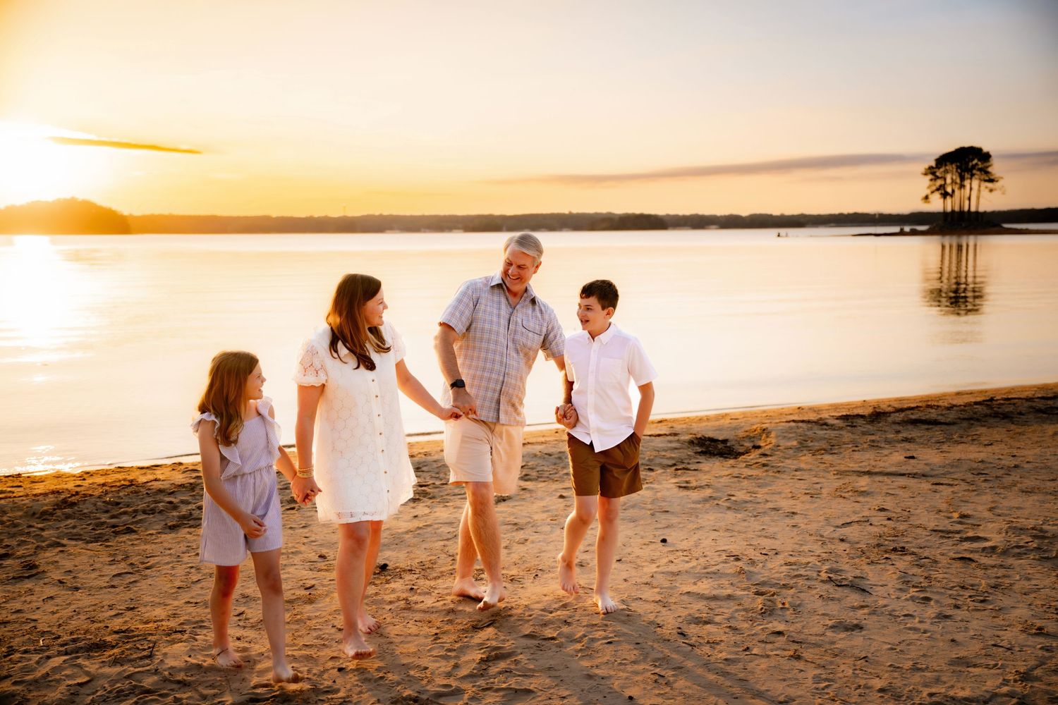 Family of four holding hands on sandy beach at golden sunset, with calm water and warm orange sky in background.