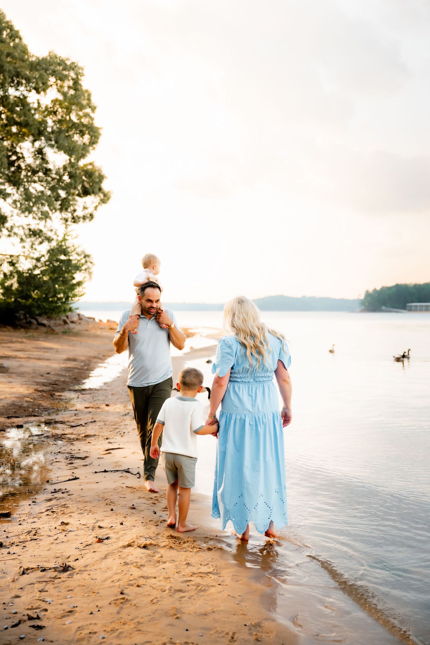 Family walking along sandy beach at sunset, father carrying toddler on shoulders, mother in blue dress holding child's hand.