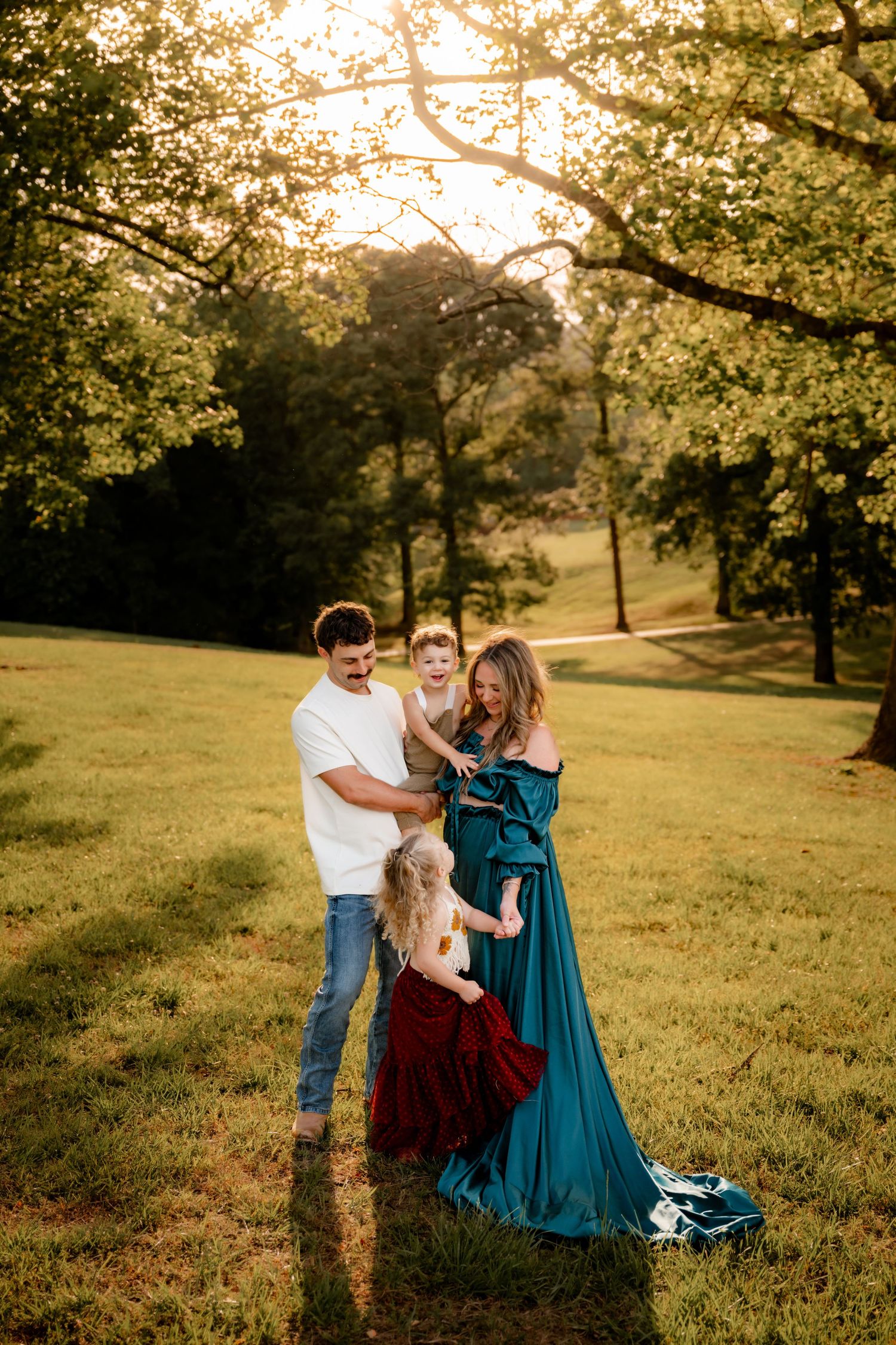 Family of four poses in golden hour sunlight in a lush green park, mother in teal gown, children in festive attire.