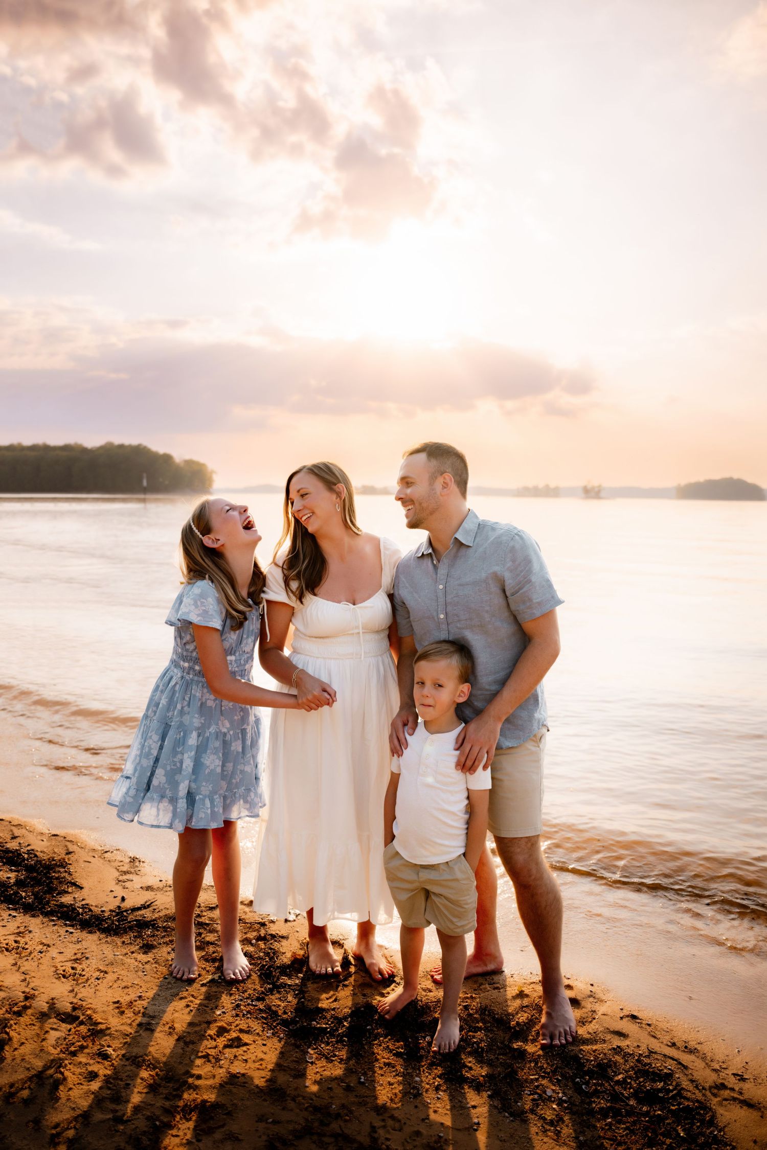 Family of four smiling together on a sandy beach at golden sunset, mother in white dress, daughter in blue floral dress.
