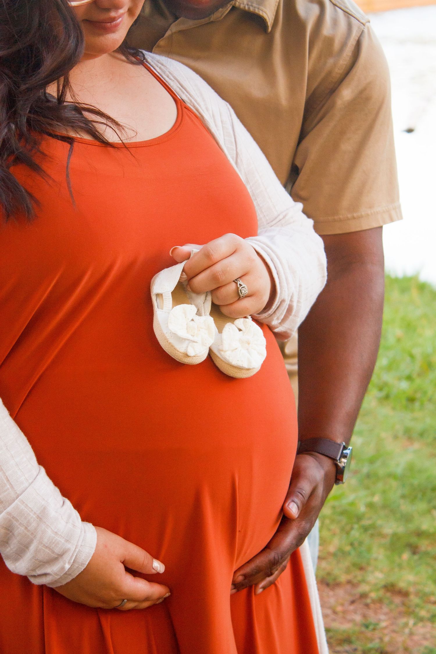 A close-up of tiny baby shoes held against a pregnant belly in an orange dress.