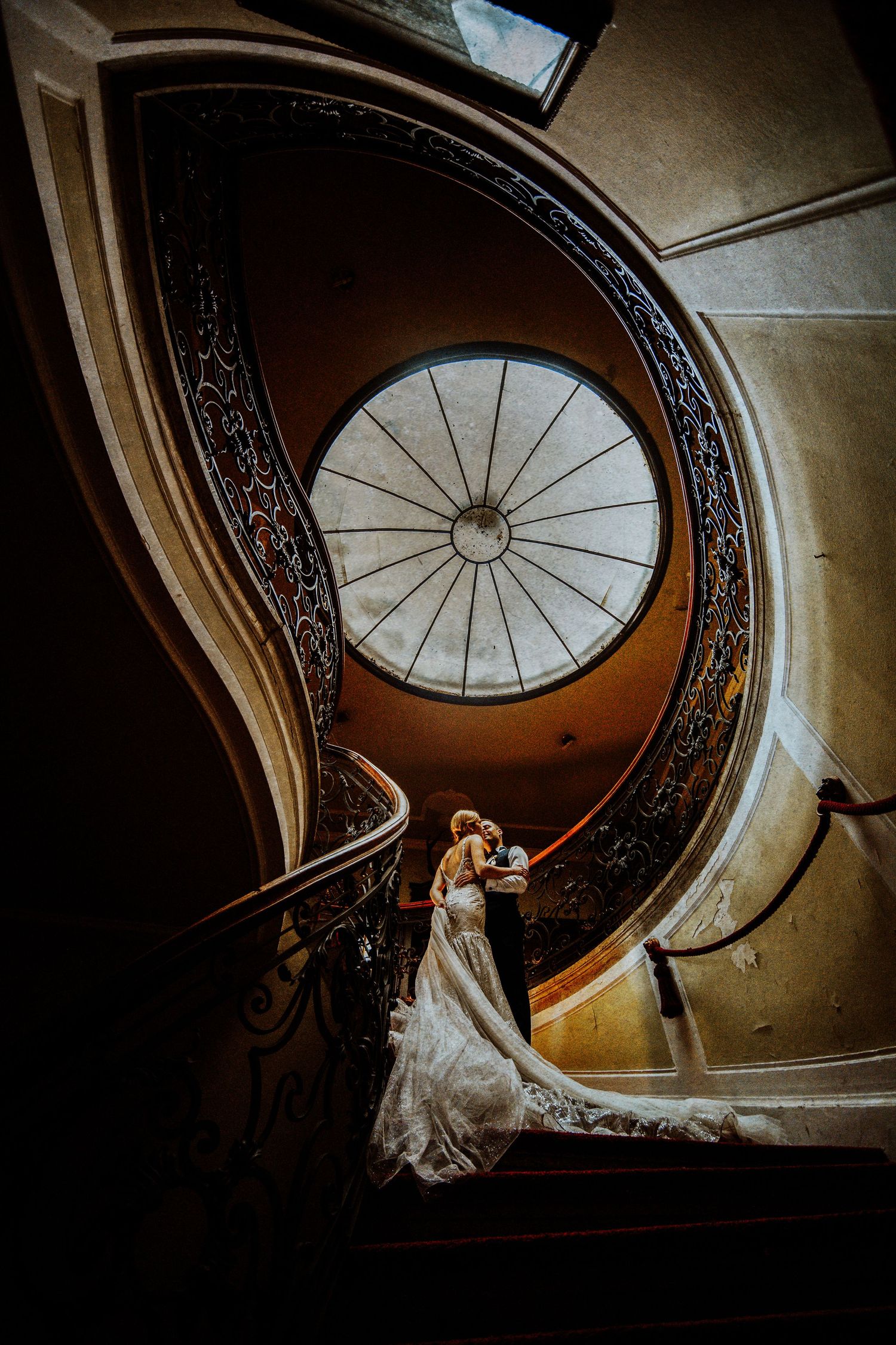 A couple in wedding attire embraces on an elegant spiral staircase beneath a domed skylight with ornate railings.