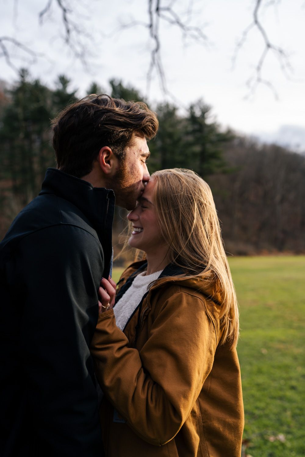 A couple embraces outdoors in a grassy field on an autumn day wearing cozy jackets.
