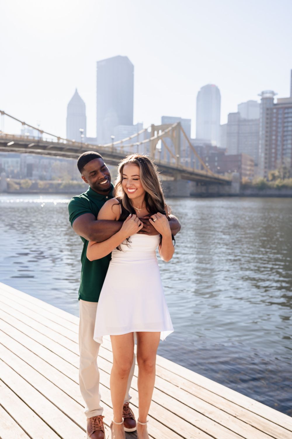 A couple embraces on a wooden pier overlooking a city waterfront with bridges and skyscrapers visible.