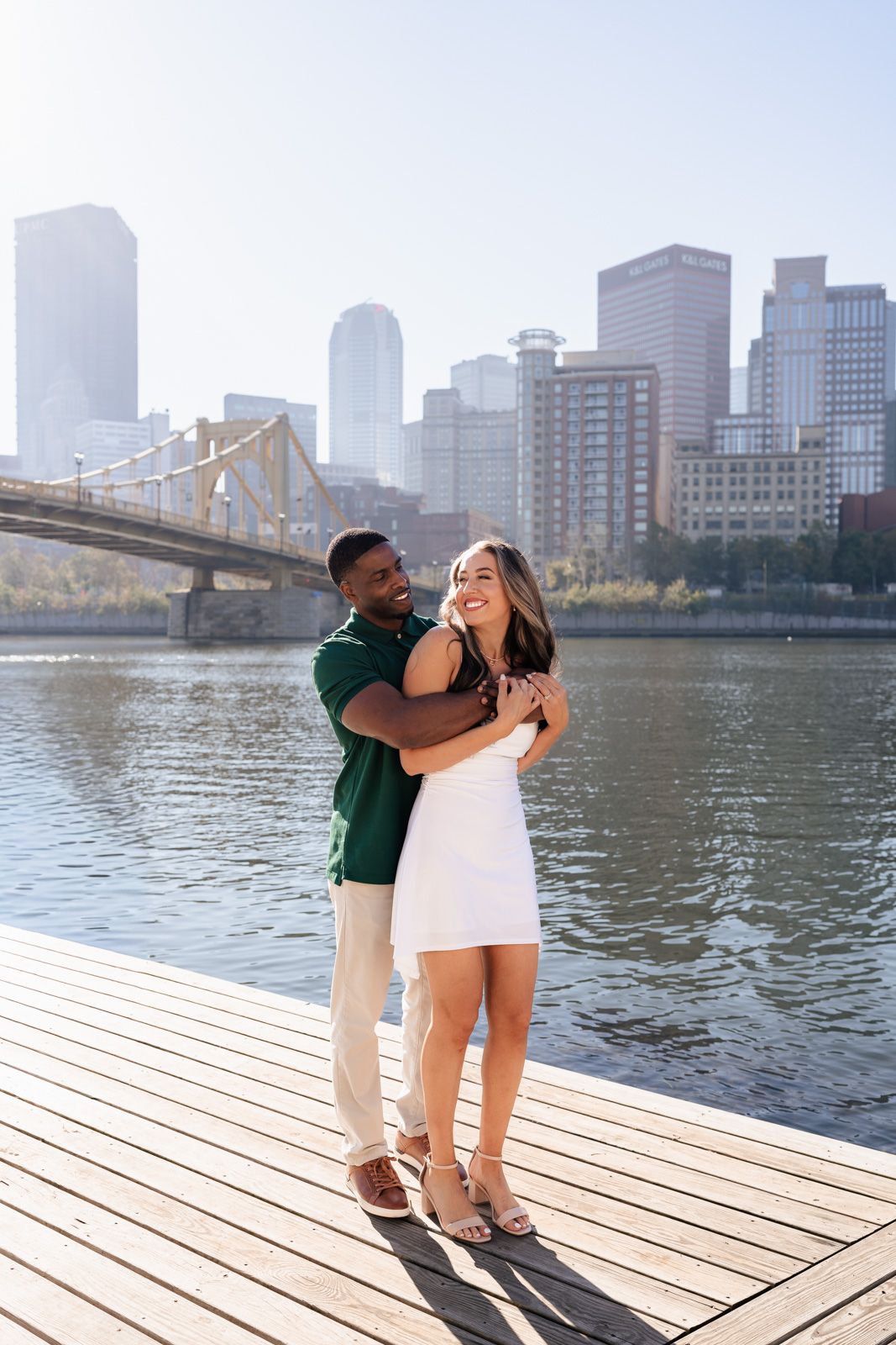 A couple shares a romantic moment on a waterfront dock with Pittsburgh's skyline and bridges in the background.