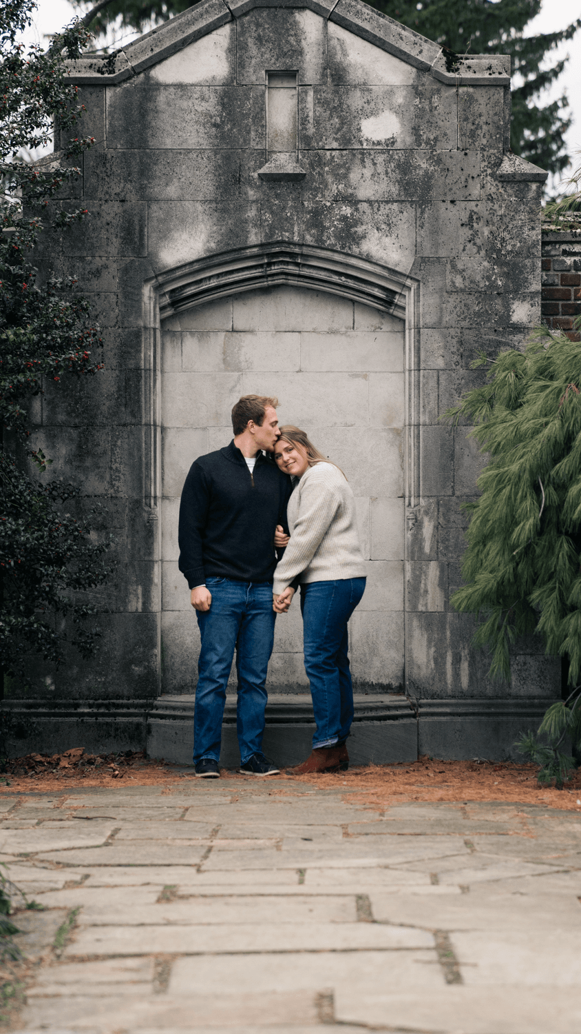 A couple shares an intimate moment against a weathered stone archway on a cloudy autumn day.