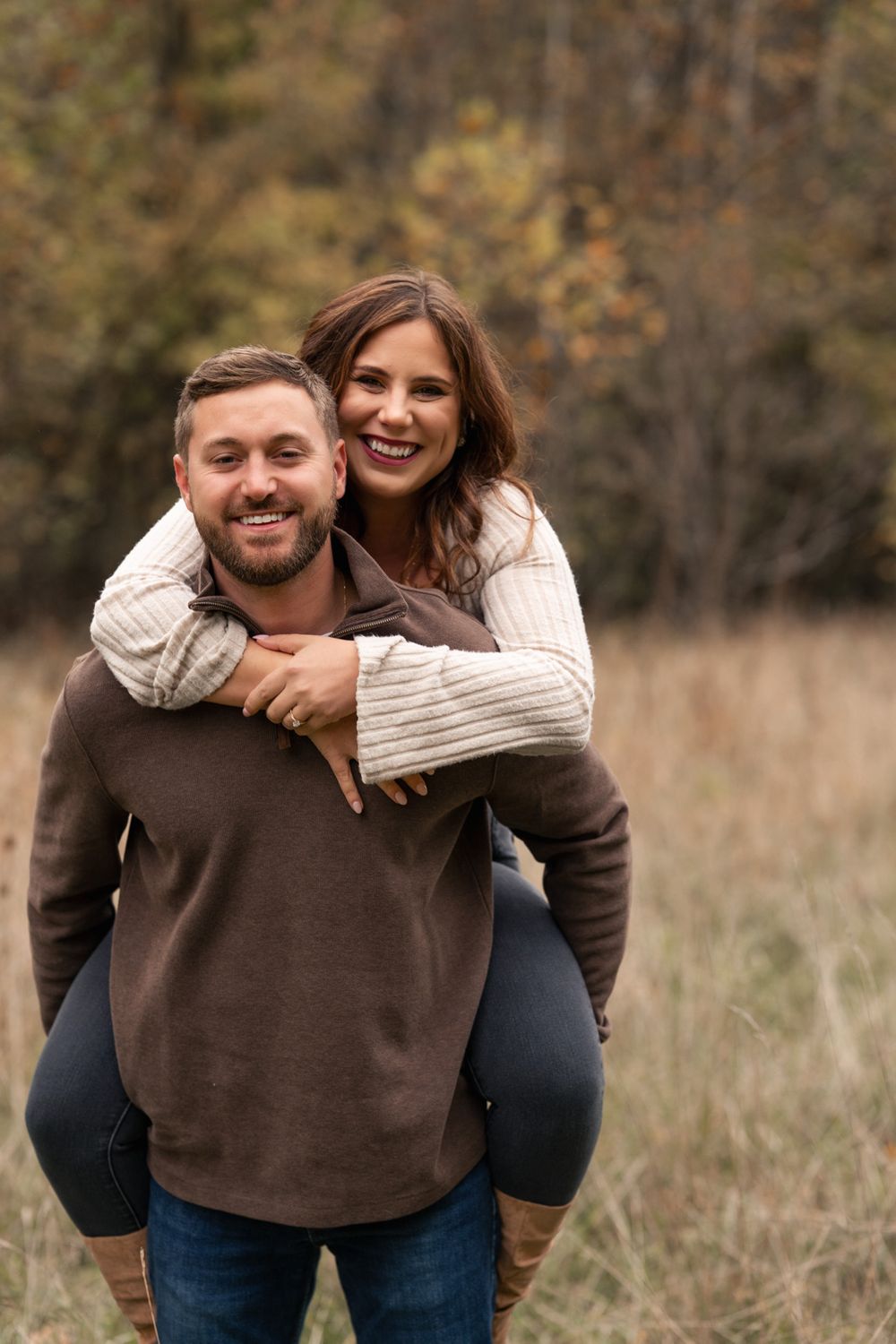A couple shares playful piggyback moments in a field during autumn with warm brown tones and natural lighting.
