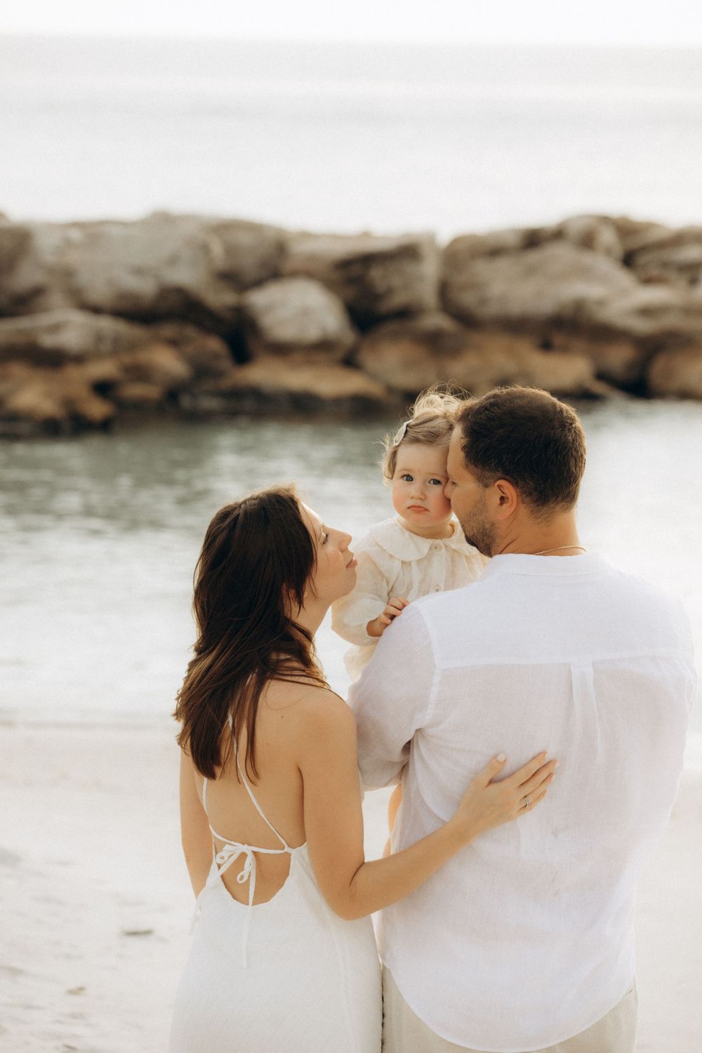 A tender family moment captured on a rocky beach as they embrace in coordinated white outfits.