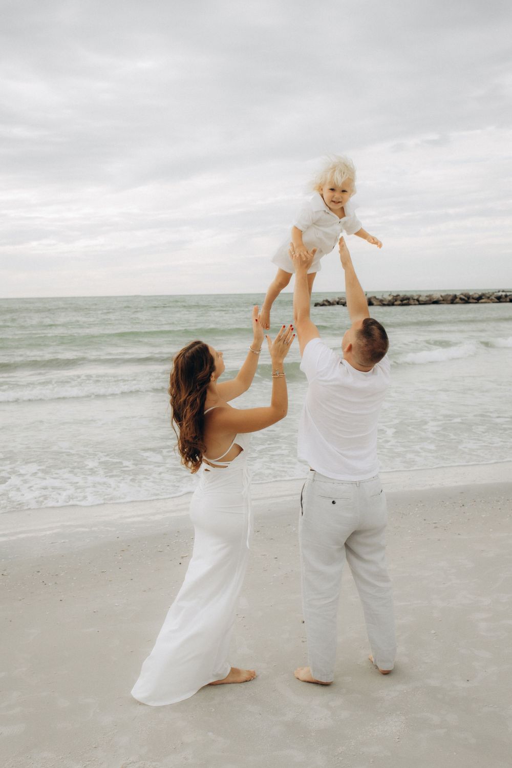 Family in white clothes playing and lifting child in the air on beach.