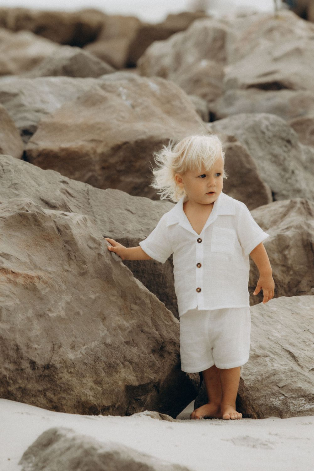Young child in white button-down outfit exploring rocky beach terrain.