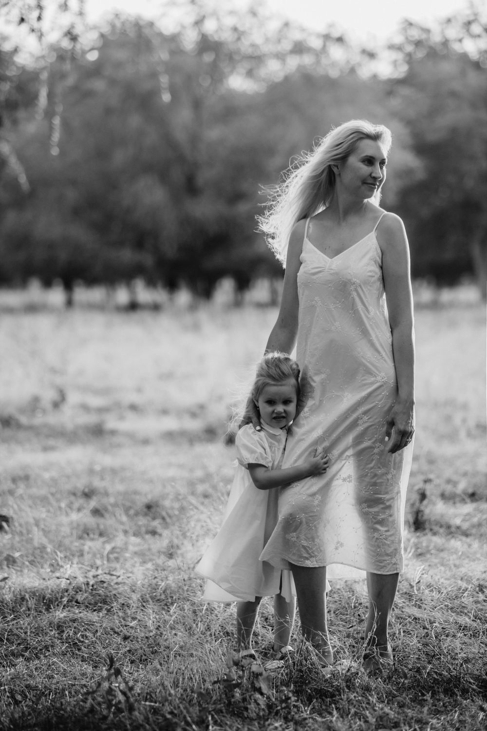 Mother and child in white dresses embracing in sunny field with trees.