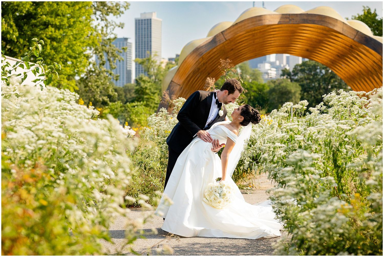 A romantic wedding moment captured under a wooden arch surrounded by white wildflowers in an urban garden.