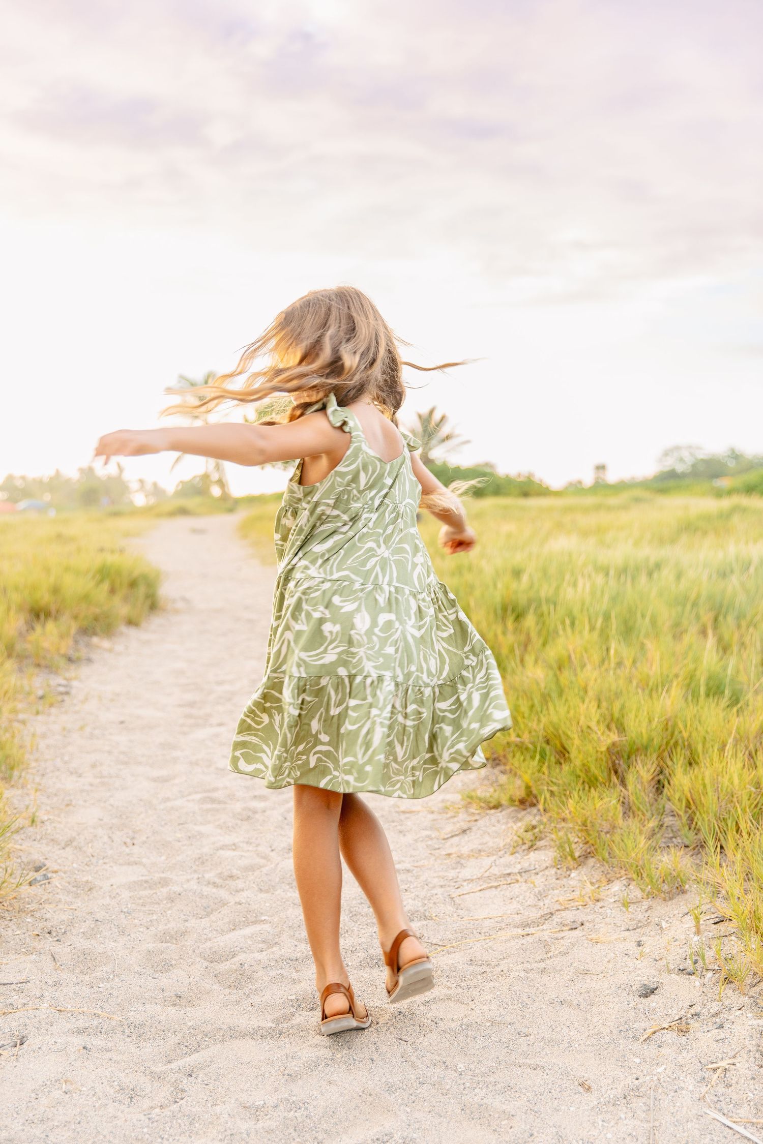 Young girl in a green floral dress twirls and dances on a sandy path through grassy fields at sunset.