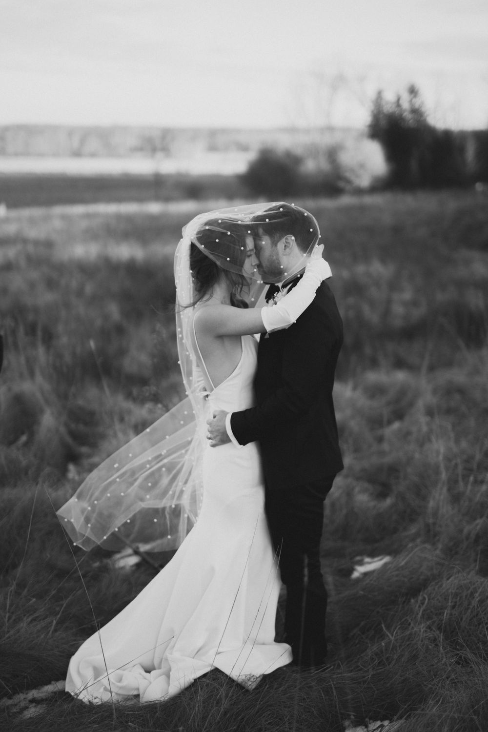 Black and white photo of newlyweds sharing intimate moment in open field, bride's pearl veil draped elegantly.