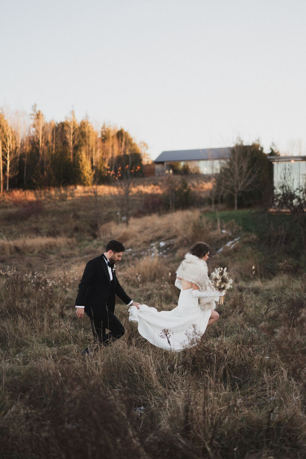 Groom helping bride lift her wedding dress train while walking through rustic countryside field at golden hour.