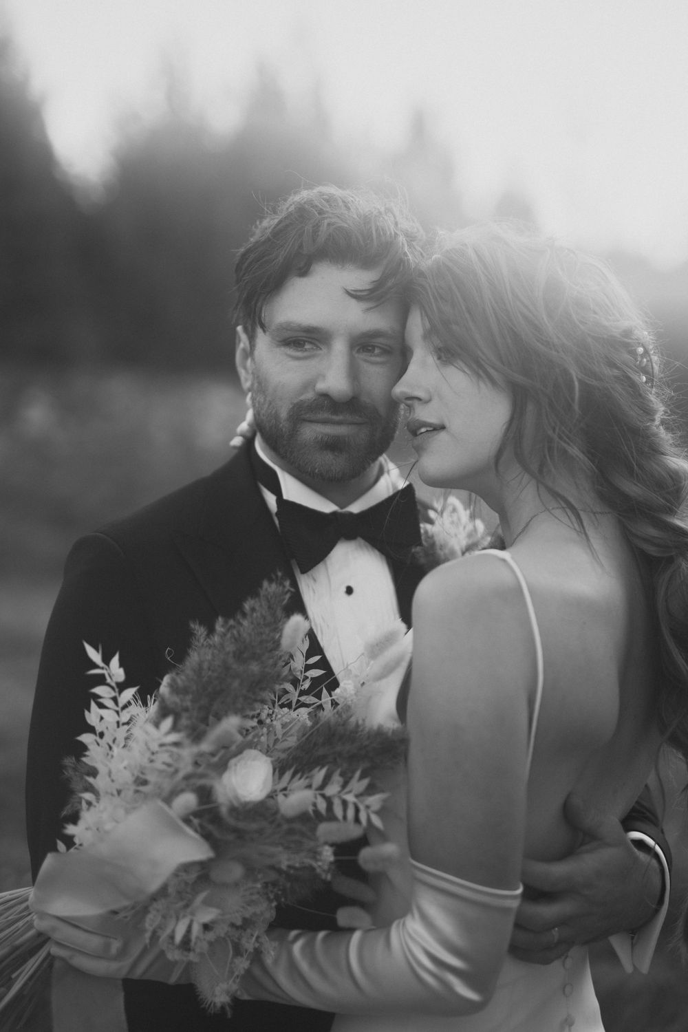 Black and white portrait of groom holding bouquet as bride leans into him tenderly in misty outdoor setting.