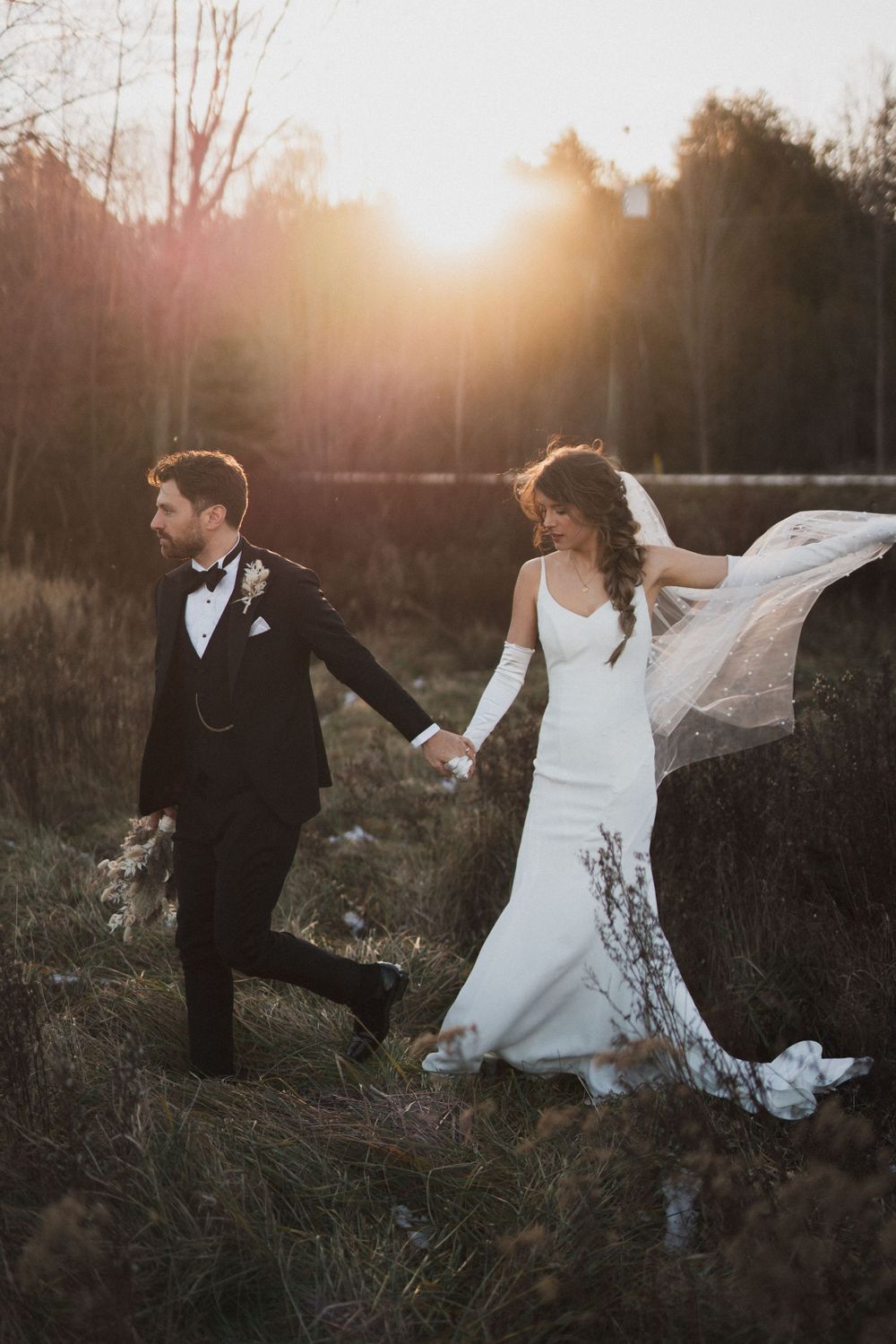 Bride and groom hold hands walking through golden sunset field, her lace veil flowing in the wind.