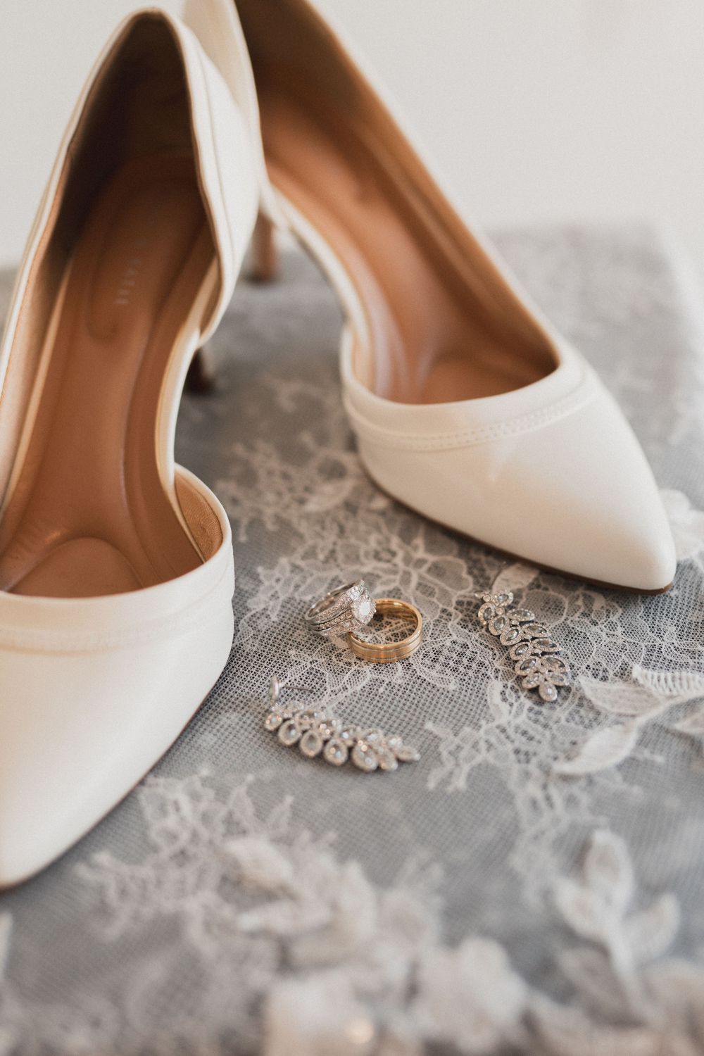 White bridal heels with gold wedding rings and crystal earrings on lace fabric detail.