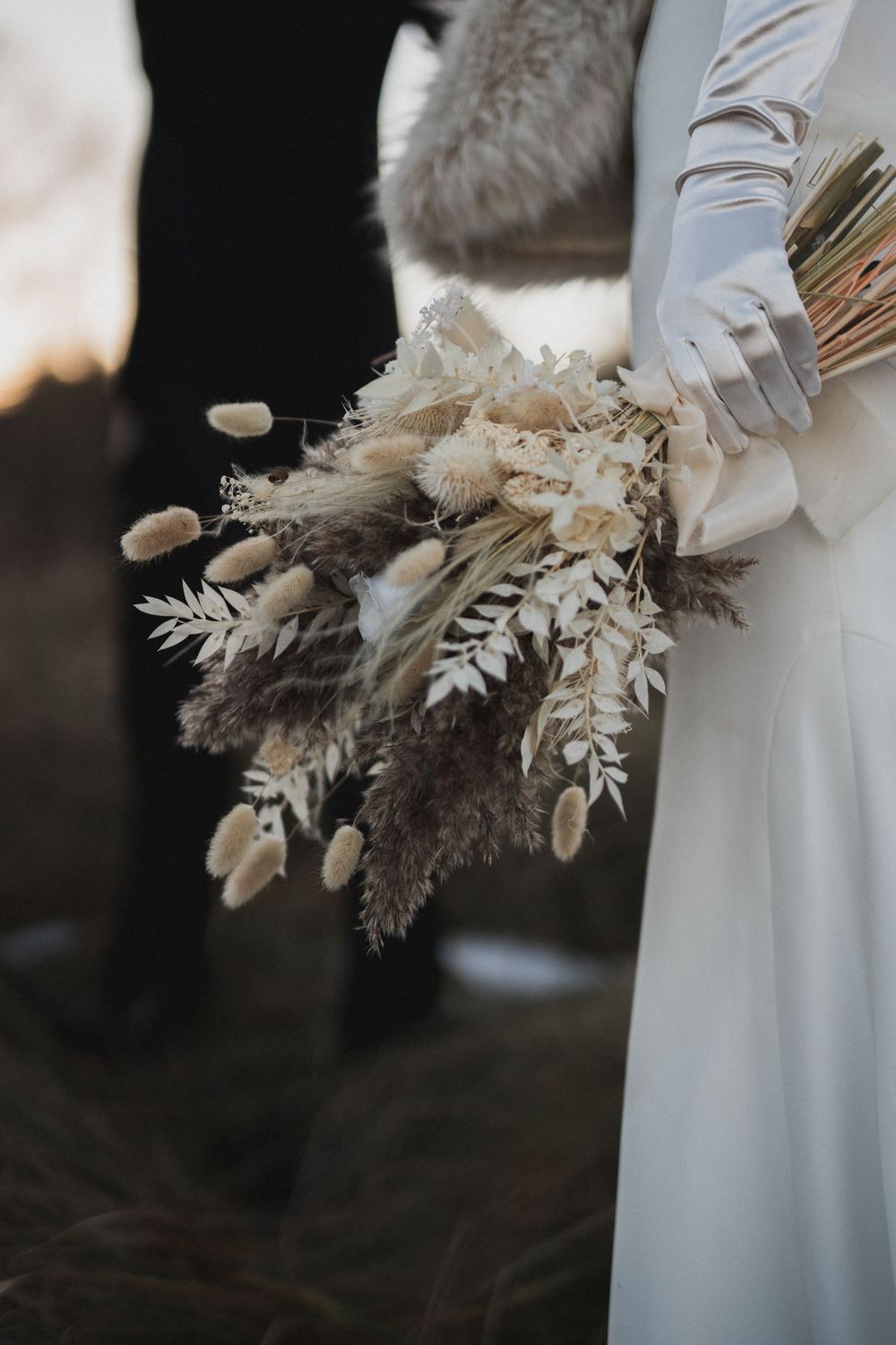 Bride holding boho dried flower bouquet with pampas grass and bunny tail stems, wearing white satin gloves.
