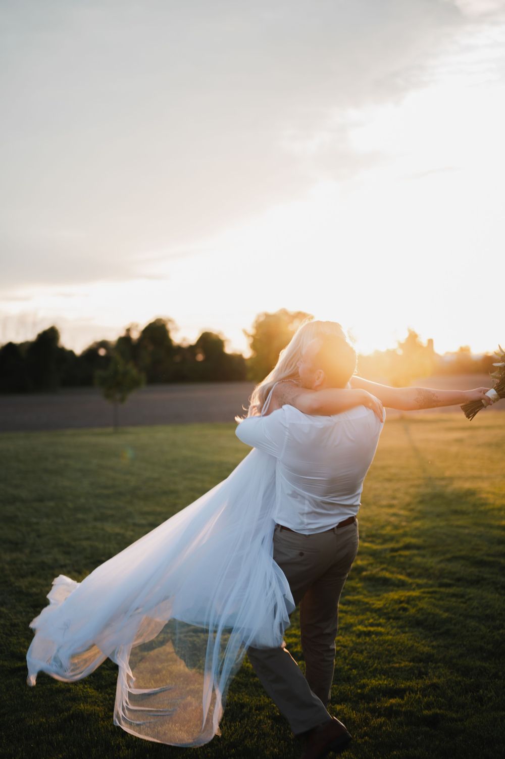 Groom lifting bride in wedding dress at golden sunset in an open field.