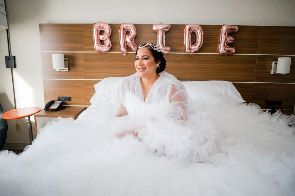 Fluffy white wedding dress surrounds a bride on a hotel bed with rose gold BRIDE balloons on the wall.