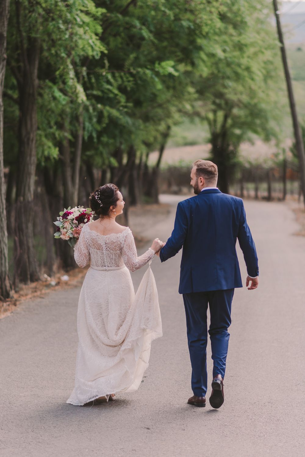 Couple walking hand in hand down a tree-lined path in wedding attire.